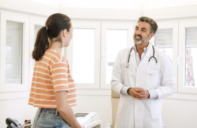 Happy mature male doctor talking to young female patient while standing during consultation in clinic